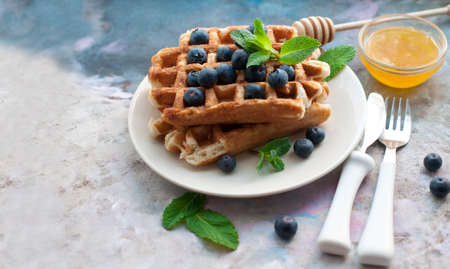 Belgian waffles with blueberries, honey and mint on a white plate. Breakfast with a cup of tea with a white teapot and sweet waffles. Knife and fork with white handlesの写真素材