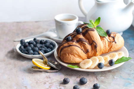 Croissant with chocolate, blueberries, bananas, and mint on a dark brown background. Breakfast with a croissant and a cup of teaの写真素材