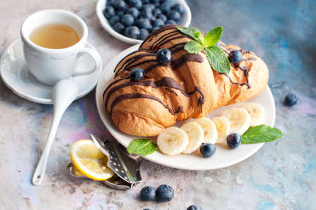 Croissant with chocolate, blueberries, bananas, and mint on a light gray background. Breakfast with a croissant and a cup of teaの写真素材