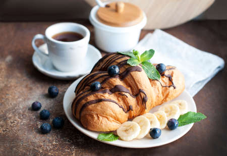 Croissant with chocolate, blueberries, bananas, and mint on a dark brown background. Breakfast with a croissant and a cup of coffeeの写真素材