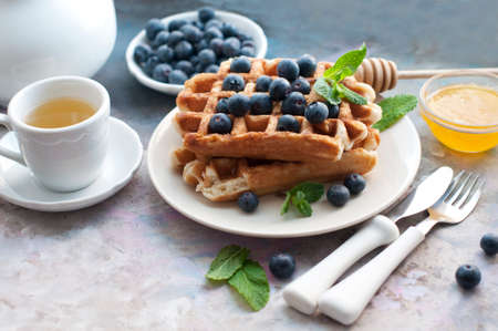 Belgian waffles with blueberries, honey and mint on a white plate. Breakfast with a cup of tea with a white teapot and sweet waffles. Knife and fork with white handlesの写真素材