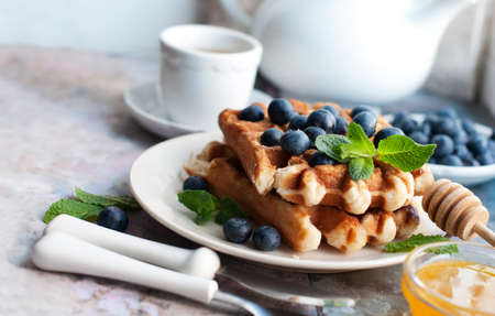Belgian waffles with blueberries, honey and mint on a white plate. Breakfast with a cup of tea with a white teapot and sweet waffles. Knife and fork with white handlesの写真素材