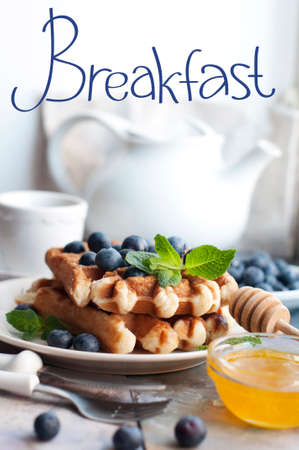 Belgian waffles with blueberries, honey and mint on a white plate. Breakfast with a cup of tea with a white teapot and sweet waffles. Knife and fork with white handlesの写真素材