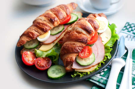 Croissant sandwich with ham, cheese, fresh salad, tomatoes, eggs and cucumber on black plate on white background. Breakfast with fresh croissant sandwich and teaの写真素材