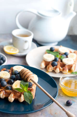 Belgian waffles with blueberries, bananas, honey and mint on a white and blue plates. Breakfast with a cup of tea with a white teapot and sweet waffles. Knife and fork with white handlesの写真素材