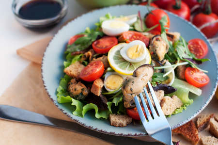 Salad with mussels, tomatoes, eggs, arugula, spinach, lemon and croutons on blue background. Seafood salad with vine sauce. Mussel on forkの写真素材