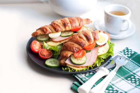 Croissant sandwiches with ham, cheese, fresh salad, tomatoes, eggs and cucumber on black plate. Breakfast with fresh croissant sandwich and cup of tea on white backgroundの写真素材