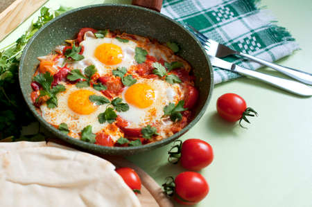 Traditional dish of israeli cuisine Shakshuka. Fried eggs with tomatoes, peppers, onions, cilantro and pita in a pan on light green backgroundの写真素材