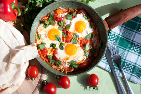 Traditional dish of israeli cuisine Shakshuka. Fried eggs with tomatoes, peppers, onions, cilantro and pita in a pan on light green background. Top viewの写真素材