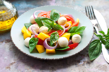 Salad with mango, mozzarella, basil, red onion, tomatoes, cream cheese and olive oil on blue plate on light gray backgroundの写真素材