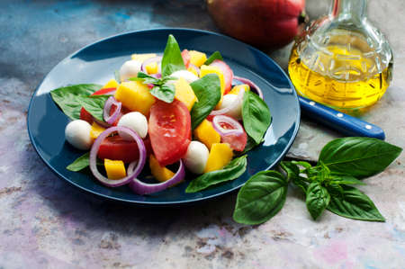 Salad with mango, mozzarella, basil, red onion, tomatoes, cream cheese and olive oil on blue plateの写真素材