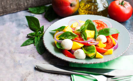 Salad with mango, mozzarella, basil, red onion, tomatoes, cream cheese and olive oil on gray plateの写真素材