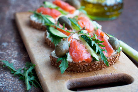 Sandwiches with trout, capers, cucumber, arugula with olive oil on wooden board on dark brown backgroundの写真素材