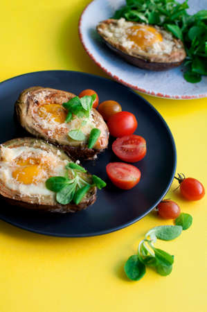 Baked avocado with egg, cheese, corn salad and tomatoes on a white and black plates on yellow background. Breakfast with avocadoの写真素材