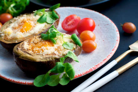 Baked avocado with egg, cheese, corn salad and tomatoes on a white plate on black background. Breakfast with avocado. Gold knife and forkの写真素材