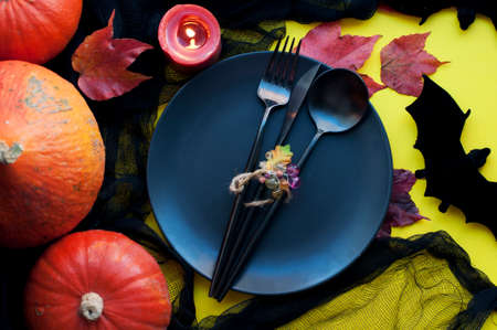 Halloween style table setting with black plate, pumpkins, bat, autumn leaves, red candle on yellow background. Black knife, black spoon, black fork. Top viewの写真素材