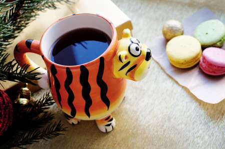 Mug in the shape of a tiger with tea and macarons on a shining gold background. Cup in the form of a tiger - a symbol of 2022. New year tea and dessert with christmas decorationsの写真素材