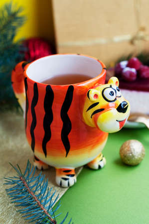 Mug in the shape of a tiger with tea and raspberry cheesecake on a green background. Cup in the form of a tiger - a symbol of 2022. New year tea and dessert with christmas decorationsの写真素材
