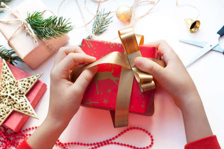 Children's hands packing a box with gifts. Boxes with gifts in red, gold and beige paper with ribbons, beads, star, fir branch. Christmas gifts with New Year's decor on a white background. Top viewの写真素材