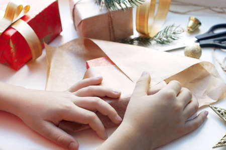 Children's hands packing a box with gifts. Boxes with gifts in red, gold and beige paper with ribbons, beads, star, fir branch. Christmas gifts with New Year's decor on a white backgroundの写真素材