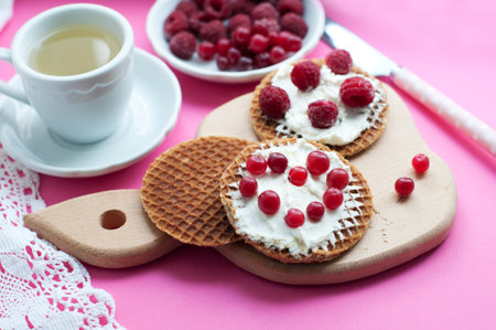 Waffles with cream cheese, cranberries and raspberries on a wooden board on a pink background. Romantic breakfast with fruits on waffles and cup of tea and white laceの写真素材