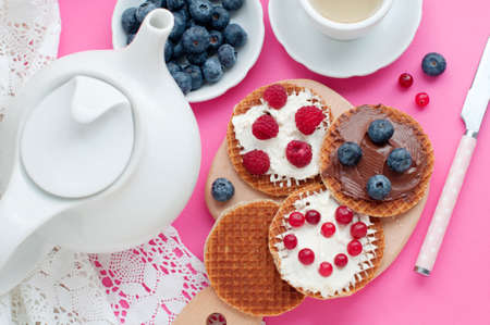 Waffles with cream cheese, cranberries, raspberries, blueberries, chocolate on a wooden board on a pink background. Romantic breakfast with fruits on waffles, teapot, cup of tea, white lace. top viewの写真素材