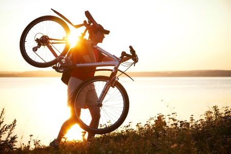 young cyclist is on the shoulder of his bike on the field at sunsetの写真素材