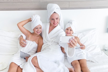 two little girl with mom in towel taking photo on phone after SPA procedures on bedの写真素材