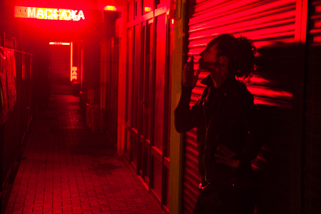 Smoking woman leaned against the wall at the red light street at nightの写真素材