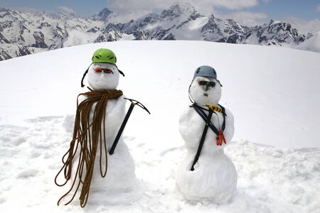 Two funny snowmen with climbing equipment stand on the background of beautiful snowy mountainsの写真素材