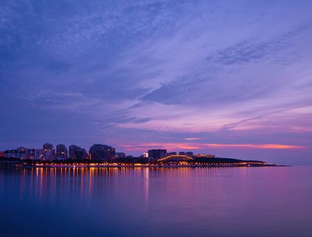 amazing colorful sunset on the sea coast with reflection of city lights in the waterの写真素材