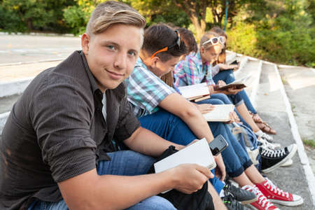 group of young students with books and gadgets sit on the steps in the parkの写真素材