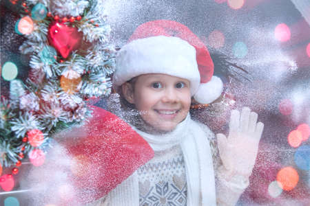 little girl in santa hat and with gifts looks into the window from winter streetの写真素材