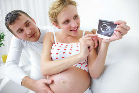 Young smiling couple holding and looking at an ultrasound scan of their expecting baby at homeの写真素材
