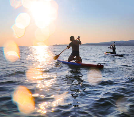 young people riding on sup surfing in the sea at sunsetの写真素材