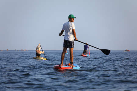 young men riding on sup surfing in the sea at sunsetの写真素材