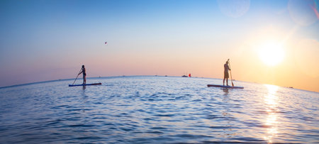 young people riding on sup surfing in the sea at sunsetの写真素材
