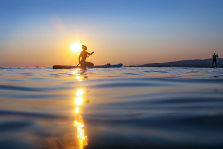 young girl riding on sup surfing in the sea at sunsetの写真素材