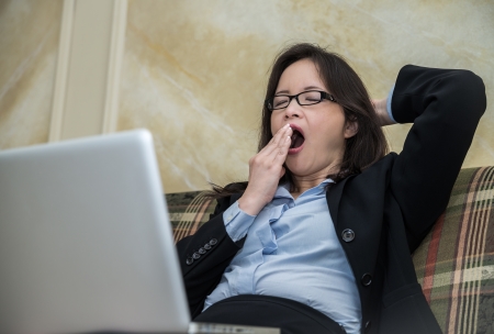 Woman in business suit feeling tired and yawning on sofa with labtopの写真素材