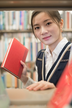 Pretty young girl picking out a red book from a pile of books on a bookshelf in libraryの写真素材