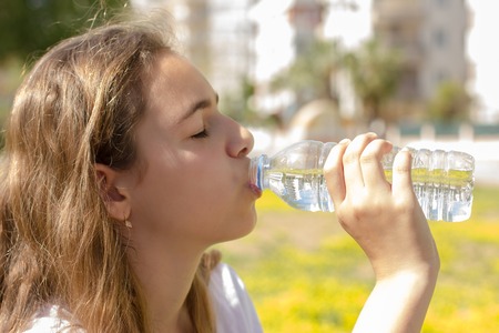 Beautiful teen girl drinks clean water from a plastic bottle on hot summer dayの写真素材