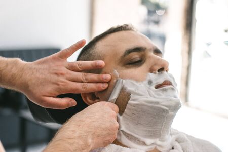 Hairdresser shaves a beard to a man with a razor in a barbershop