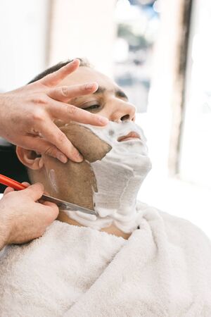 Hairdresser shaves a beard to a man with a razor in a barbershopの写真素材