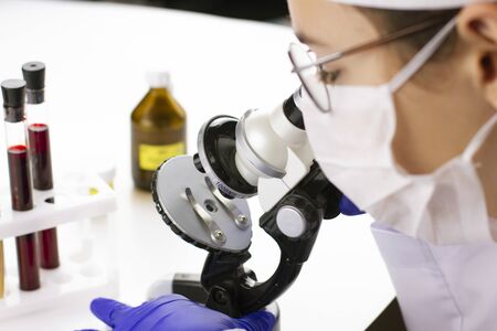 Doctor in a protective mask and gloves researching blood through microscope for coronavirus...