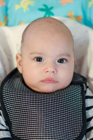 Baby in an apron sits in a highchair and waits to be fed. Serious baby looking to cameraの写真素材