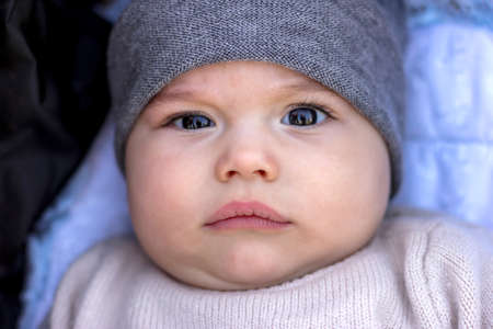 Portrait of serious baby in grey hat. Sweet little infant looks to camera. Kid with brown eyes and long lashesの写真素材