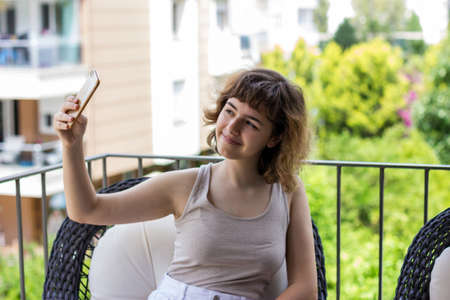 Pretty curly smiling young girl taking selfie on smartphone on the apartment balcony. Video chatting...