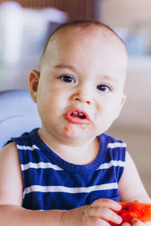 Sweet baby boy eating watermelon sitting at baby chair in restaurant. Hotel vacation with child