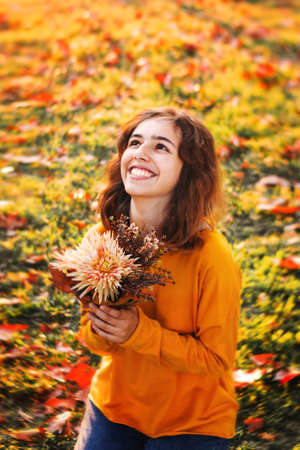 Curly young girl in yellow sweater on the grass with autumn bouquet of dry leaves and flowersの写真素材