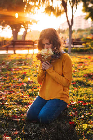 Curly young girl in yellow sweater on the grass with autumn bouquet of dry leaves and flowersの写真素材